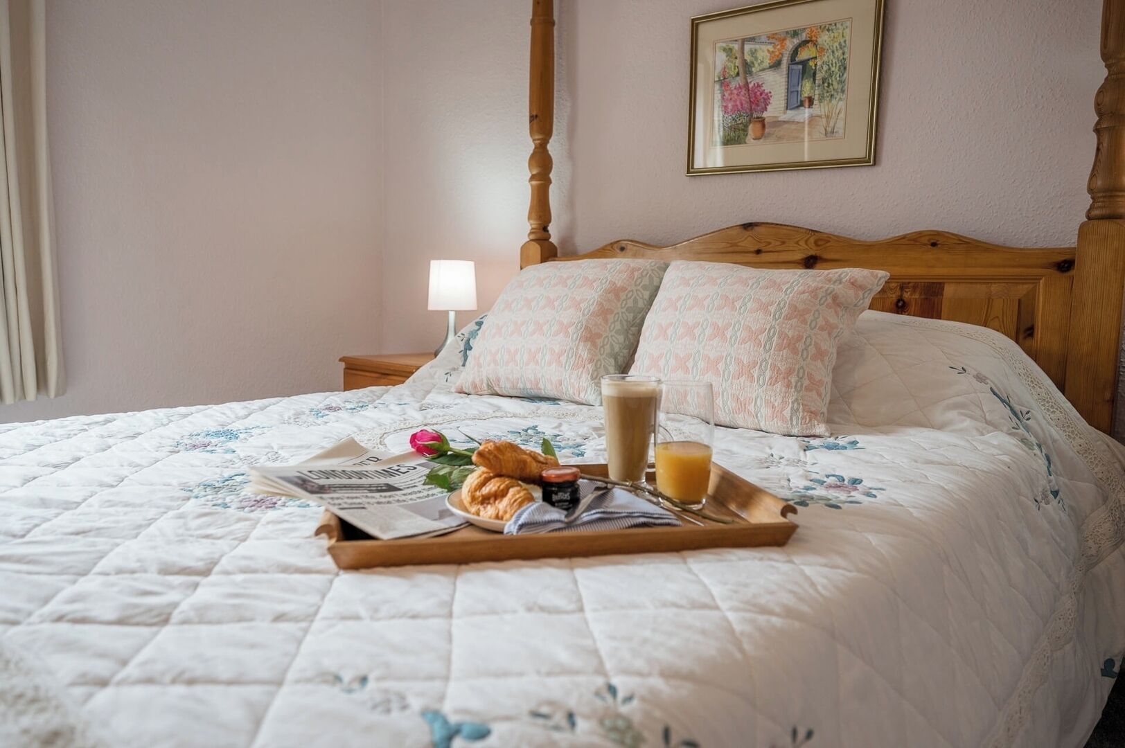 A neatly made bed with a floral quilt, a tray with croissants, a glass of orange juice, and a newspaper.