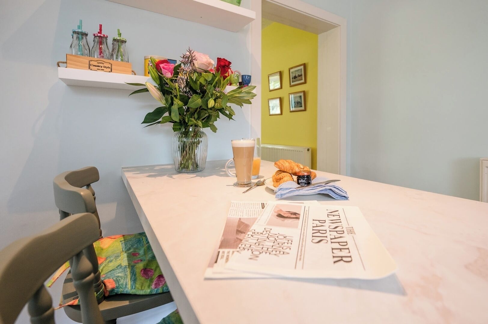 Een eettafel met een krant, een glas bier, croissants en een bloevas.