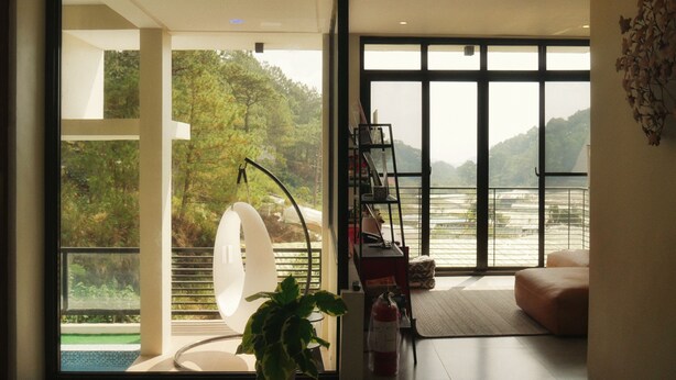 A modern interior with a large glass door leading to a balcony with a view of a forested area and a body of water. - Charming 5-bedroom Villa in Baguio with Heated Dipping Pool :Glass & Green Perch (Baguio)