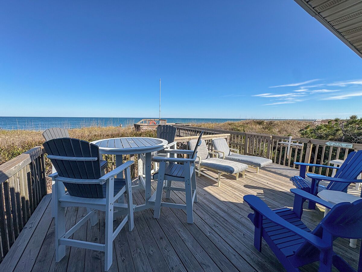 Una terraza de madera con sillas Adirondack y una mesa, con vistas a una playa y un cielo despejado.