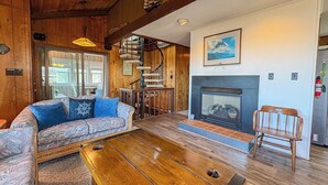 A cozy cabin interior with a wooden ceiling, a fireplace, a sofa with patterned cushions, and a dining area with a spiral staircase. - Kohler Cottage - 4 BR Oceanfront Cottage in Southern Shores w/Elevator (Southern Shores)
