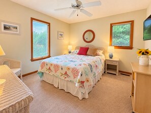 Une chambre à coucher comprenant un lit, deux tables de chevet, une chaise, un ventilateur de plafond et une fenêtre avec des stores.