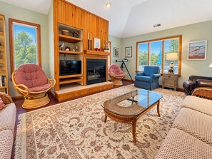 A living room with a fireplace, a television, wicker furniture, and a large window with a view.