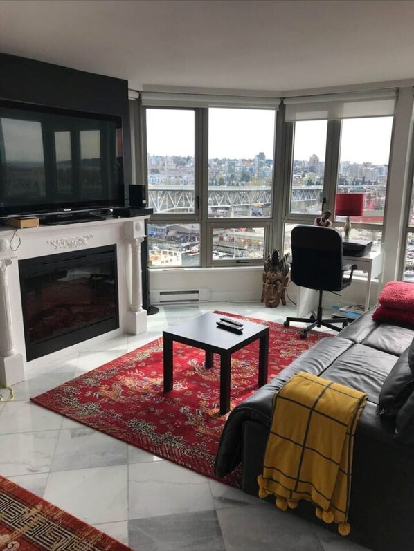 Ein Wohnzimmer mit Kamin, Fernseher, einer Couch, einem Couchtisch und großen Fenstern mit Blick auf die Stadt.