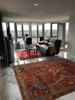 A spacious living room with a large window, a black leather sofa, a red rug, and a small table.