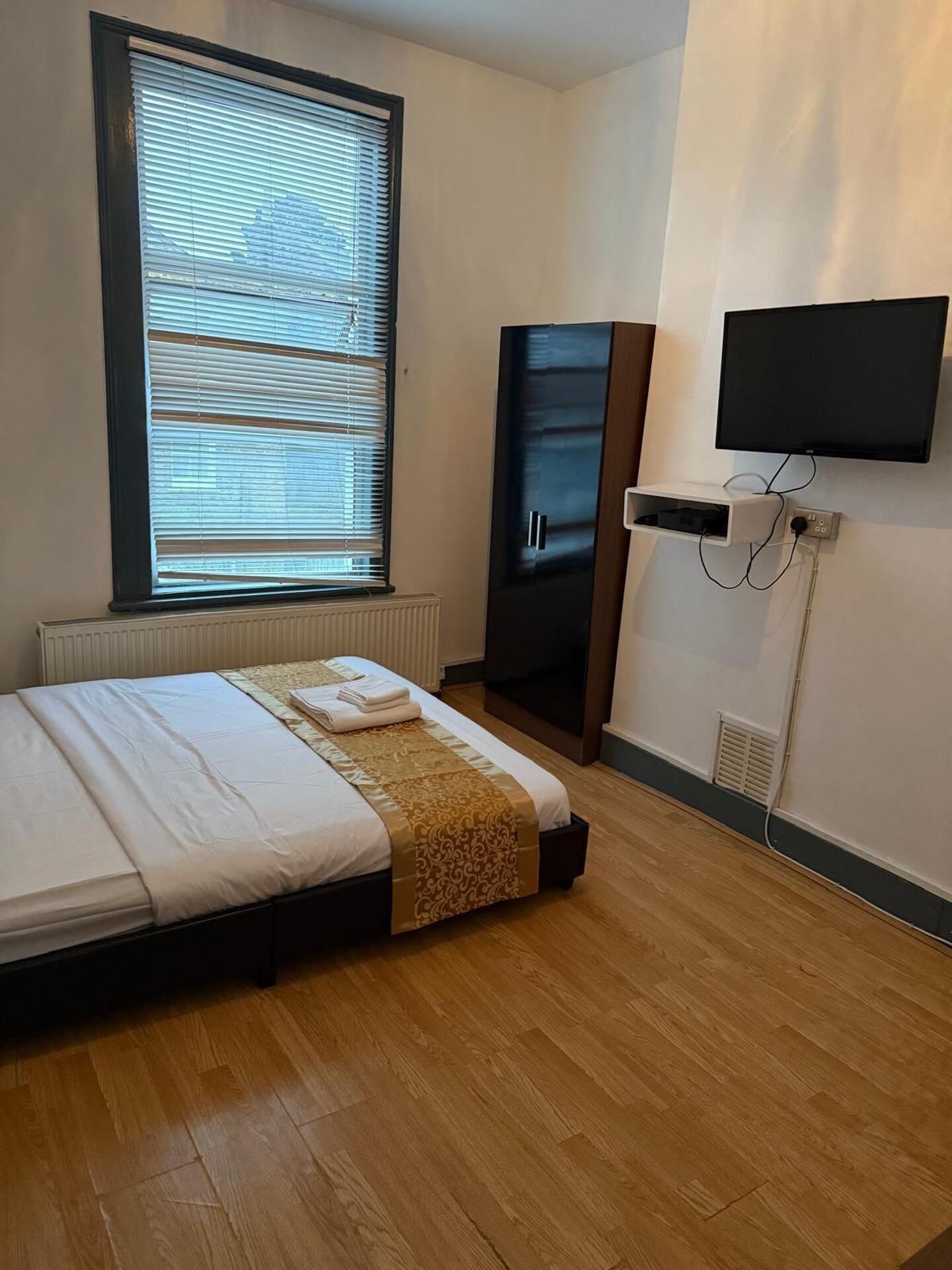 A bedroom with a bed, a large window with blinds, a wardrobe, a television mounted on the wall, and a radiator.