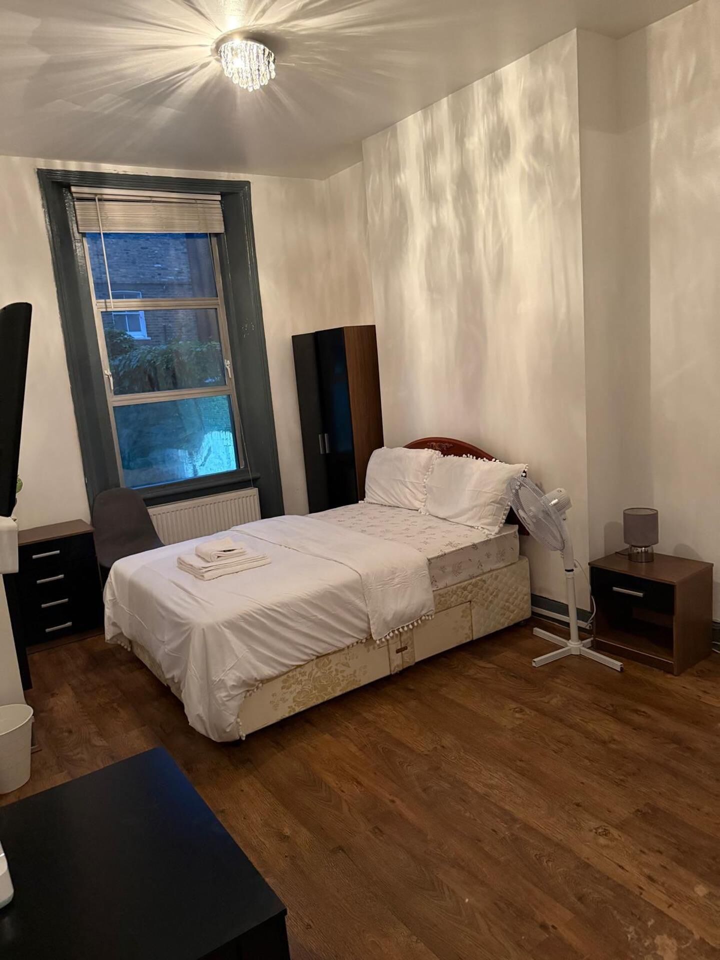 Une chambre à coucher comprenant un lit, des tables de chevet, un ventilateur sur pied, une fenêtre avec des stores et un radiateur.