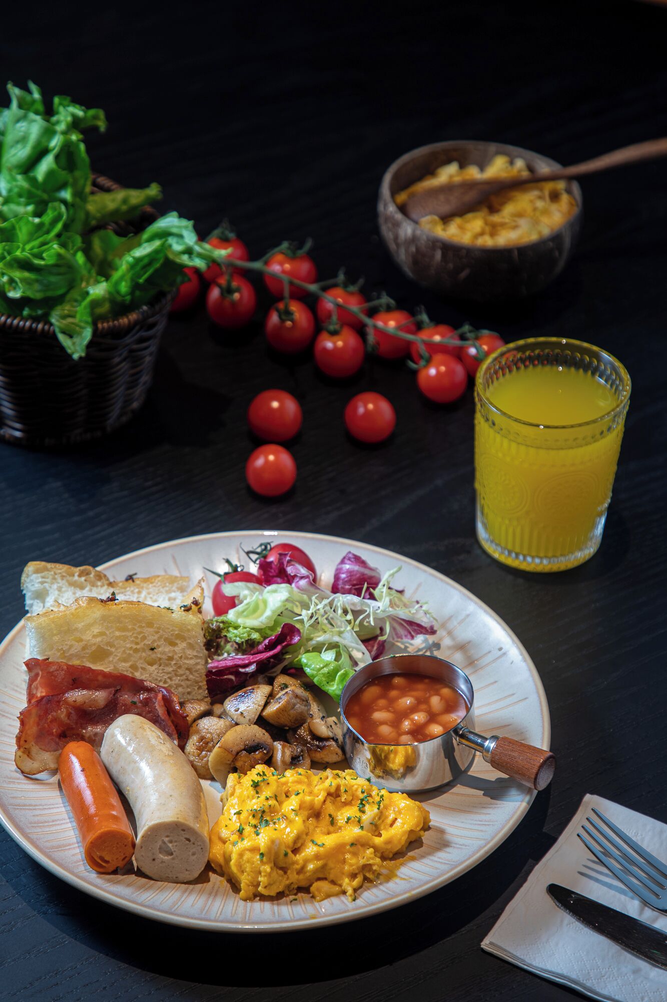 A breakfast plate with toast, bacon, sausage, scrambled eggs, mushrooms, beans, and a salad, along with a glass of orange juice and a bowl of soup.