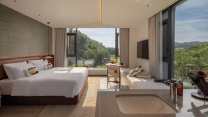 Una habitación de hotel moderna con una cama grande, un área de descanso con vistas, un ventanal amplio y un baño con lavamanos y ducha.