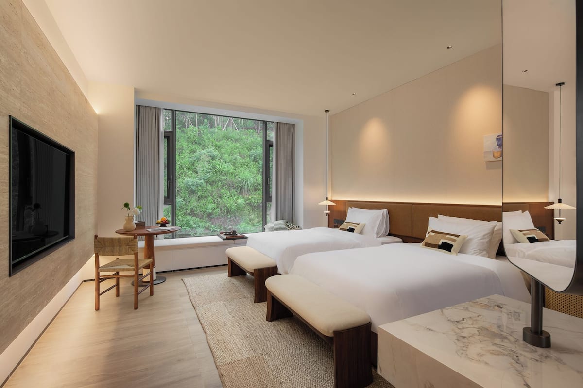 Een hotelkamer met twee bedden, een bureau en een groot raam met uitzicht op groen.