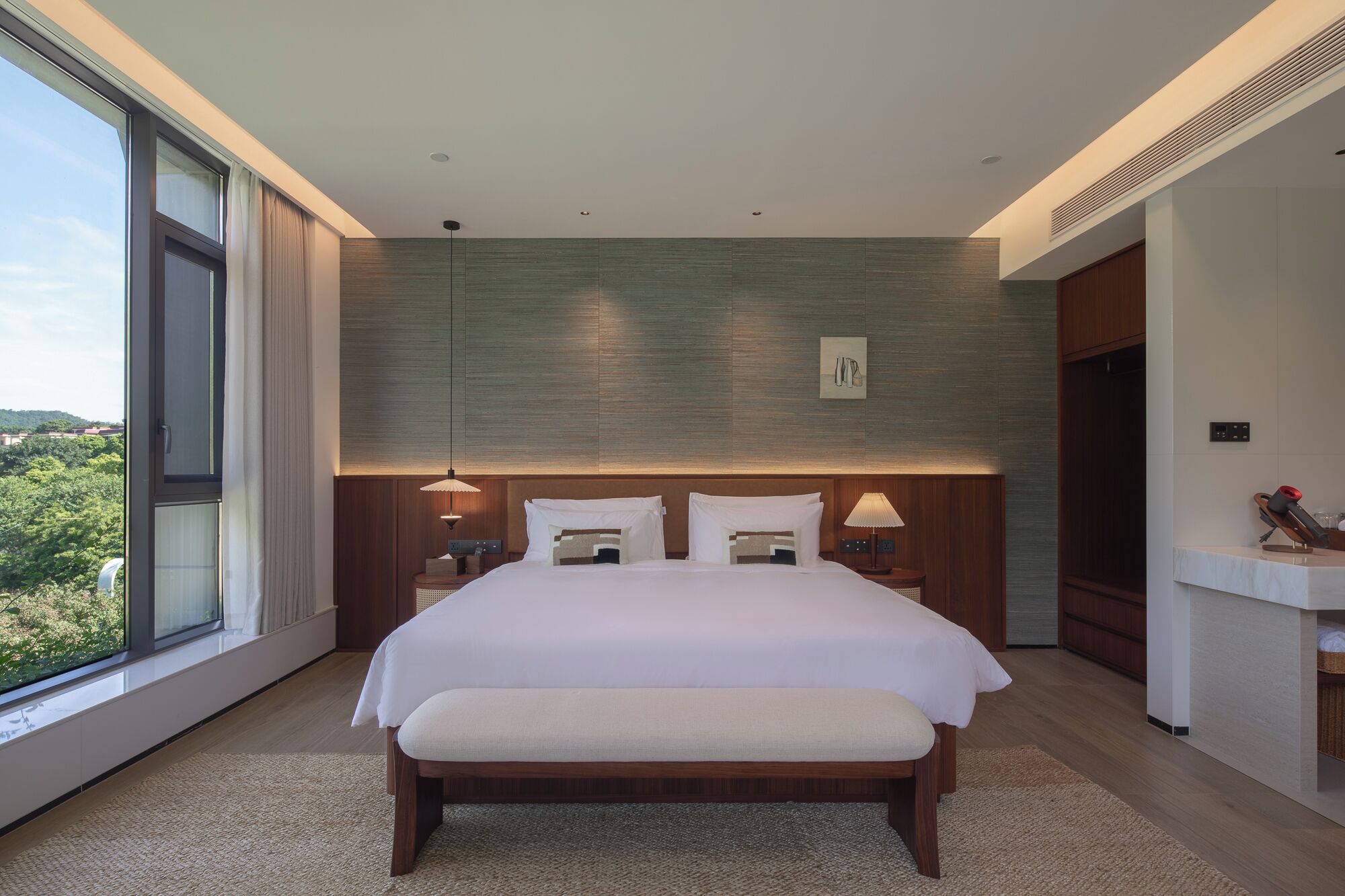 Een moderne hotelkamer met een groot bed, twee nachtkastjes, een bankje, een groot raam en uitzicht op groen.
