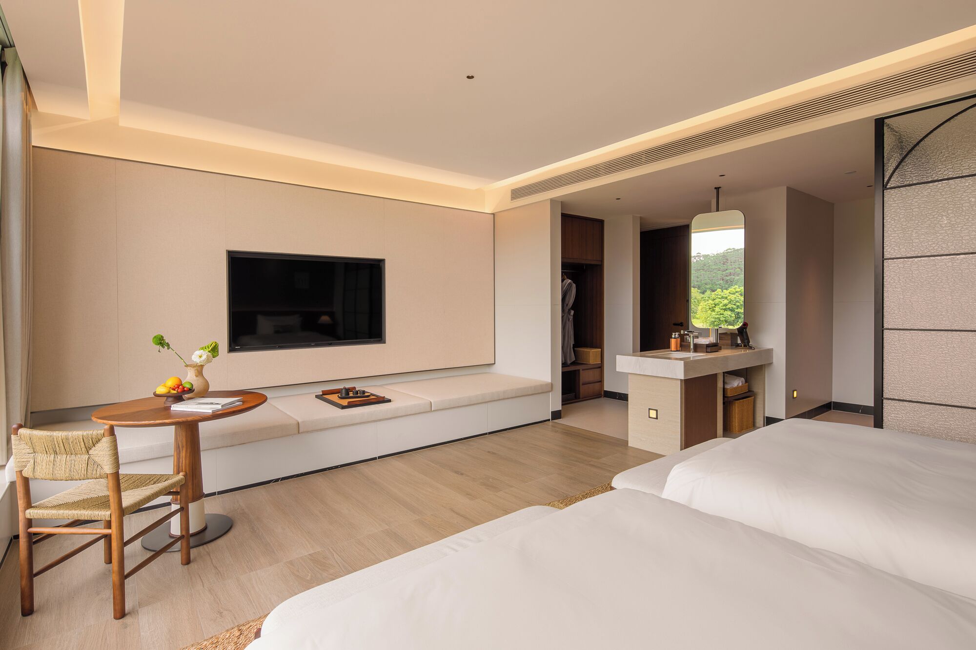 Ein modernes Hotelzimmer mit einem großen Bett, einer Sitzecke mit Fernseher, einem kleinen Tisch mit einer Vase und einem Badezimmer mit einem großen Spiegel.