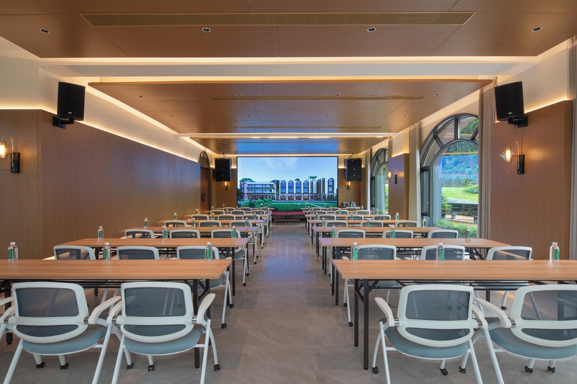 A conference room with tables and chairs, a large screen at the front, and a view of a building and greenery outside.