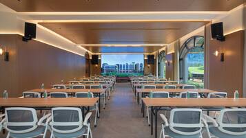 A conference room with tables and chairs, a large screen at the front, and a view of a building and greenery outside.