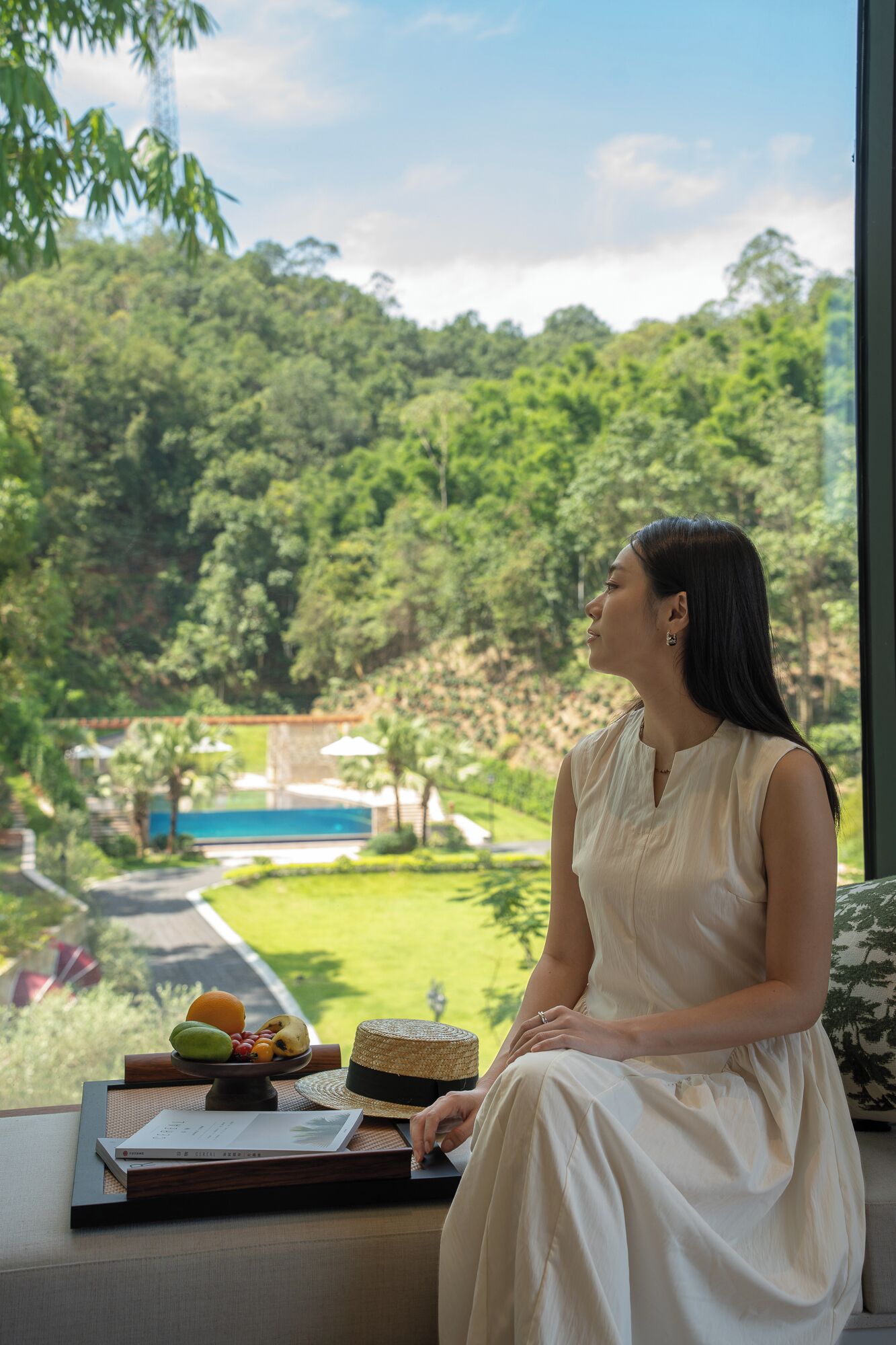 A woman seated by a window overlooking a lush garden and a pool.