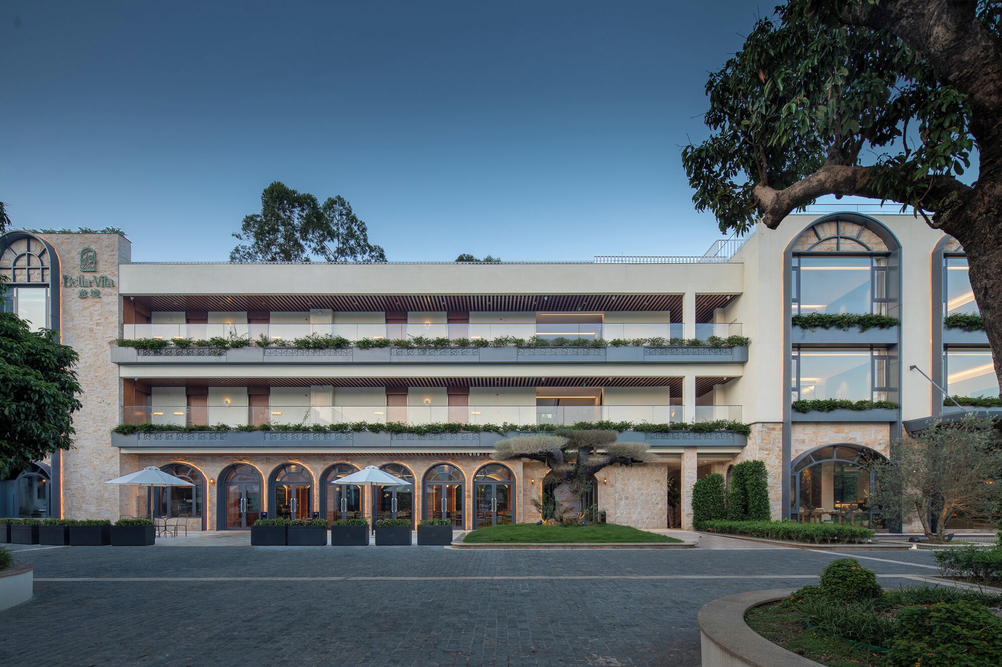 Un hôtel à plusieurs étages, doté d’un design moderne, avec des balcons agrémentés de plantes, des fenêtres en arc de cercle et une façade en pierre.