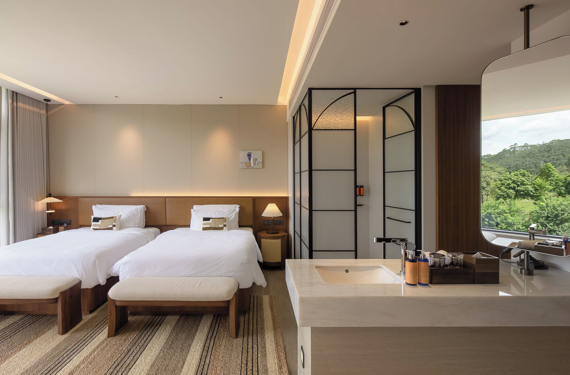 Habitación de hotel con dos camas, un espejo grande, baño con lavamanos y artículos de aseo, y vista a zonas verdes.