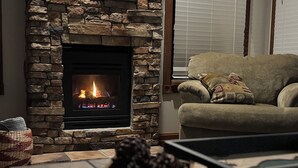 Une cheminée en pierre avec un feu de bois, un canapé agrémenté d’un coussin à motifs et un panier tressé posé sur le sol.