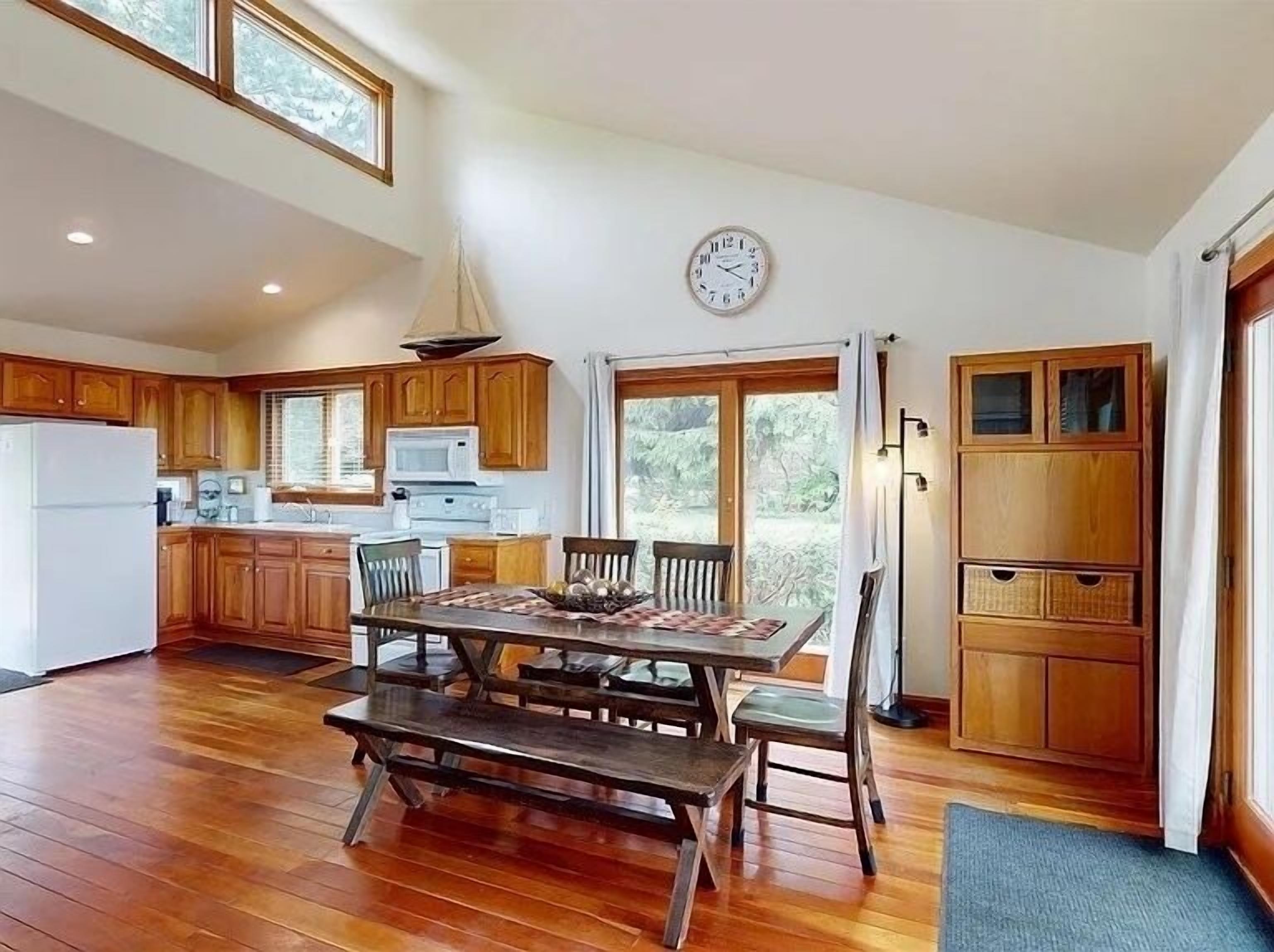 A spacious kitchen with wooden cabinets, a dining table with benches, a clock on the wall, and a view through the glass door.