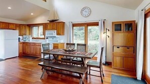 Une cuisine spacieuse avec des meubles de rangement en bois, une table à manger avec des bancs, une horloge au mur et une vue à travers la porte vitrée.