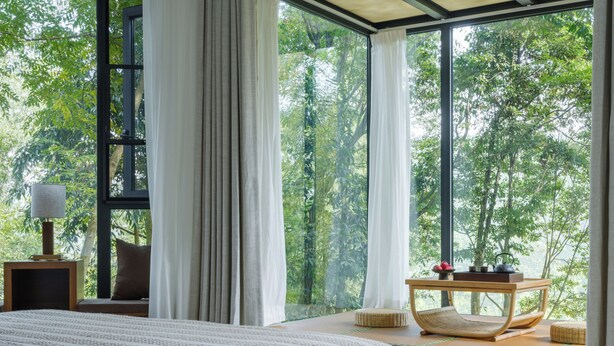 A bedroom with a large bed, a nightstand, a lamp, a chair, and a table with a view of trees through large windows. - Qingcheng Mountain Jiuxipo (Chengdu)
