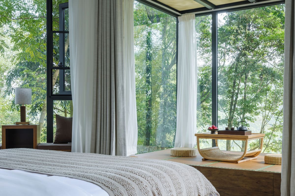 Un dormitorio con una cama grande, una mesita de noche, una lámpara, una silla y una mesa con vista a árboles a través de amplios ventanales.