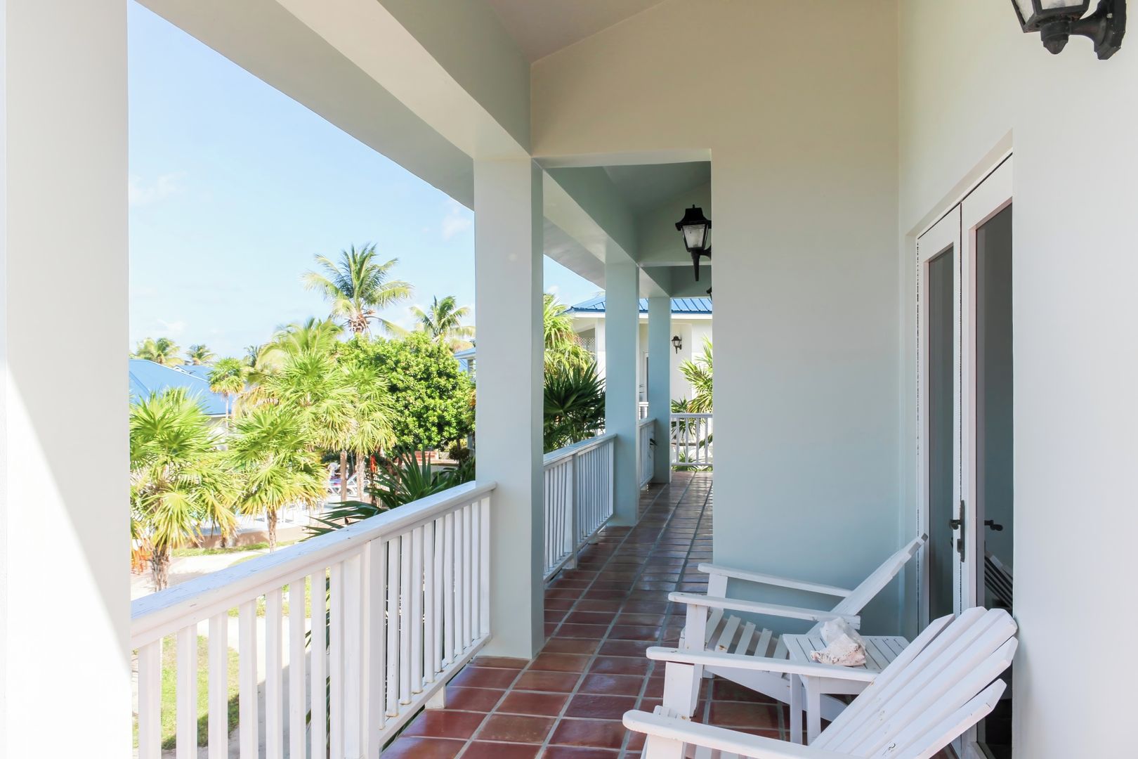 A covered balcony with white railings, a tiled floor, and a view of palm trees and a blue sky.
