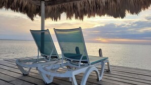 Two lounge chairs under a thatched roof, overlooking a calm sea at sunset. - 4 BDRM Sunshine Suites @ Sapphire Beach Resort (Not available)