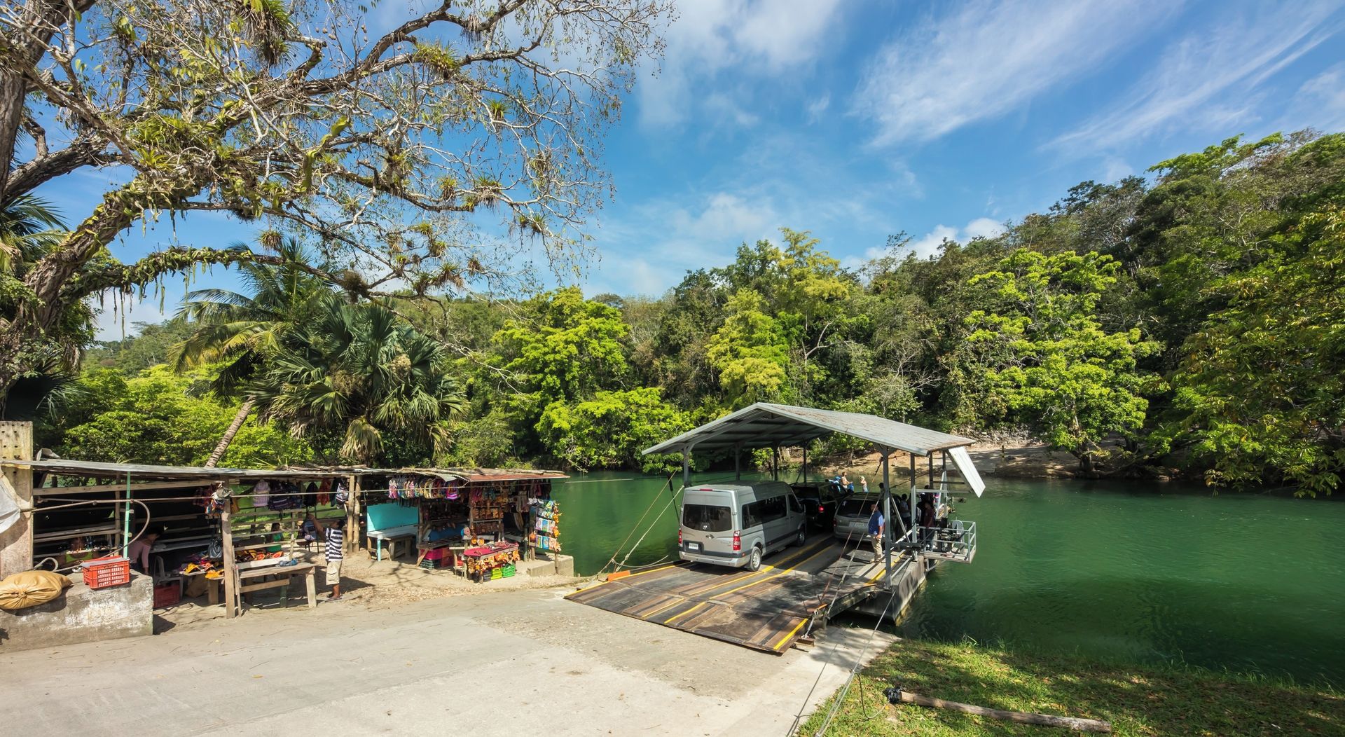 A riverside parking area with a covered vehicle bay and a small market stall selling goods.