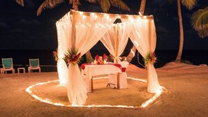 A beachside tent with a table set for two, surrounded by palm trees and string lights. - 4 BDRM Sunshine Suites @ Sapphire Beach Resort (Not available)