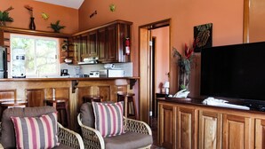 A cozy living room with wicker furniture, a flat-screen TV, and a kitchen area with wooden cabinets and a window overlooking greenery. - 4 BDRM Sunshine Suites @ Sapphire Beach Resort (Not available)