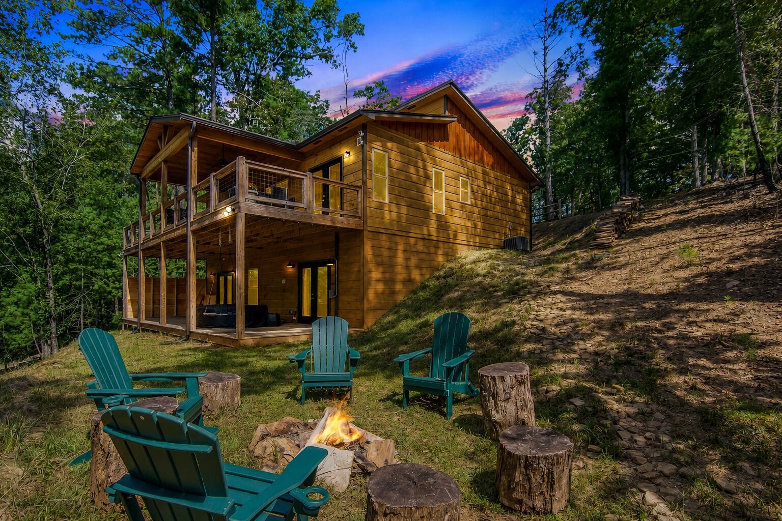 A wooden cabin with a balcony, surrounded by trees, and a fire pit with chairs.