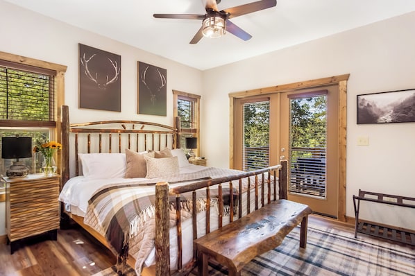 Une chambre à coucher avec un lit en bois, un banc, un ventilateur de plafond et une vue sur l’extérieur grâce à de grandes fenêtres.