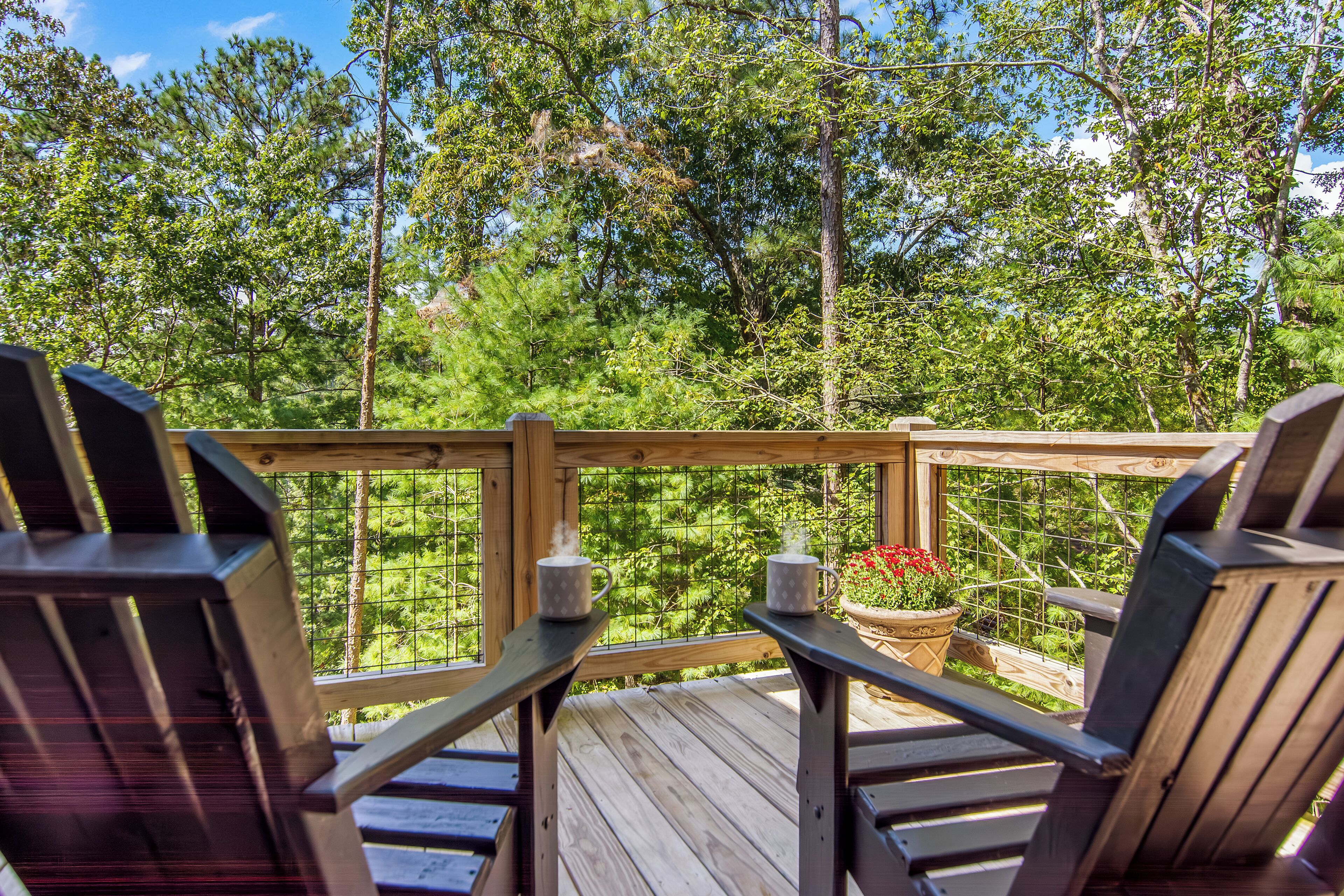 Una terraza de madera con dos sillas Adirondack y una mesa, con vista a un área boscosa.