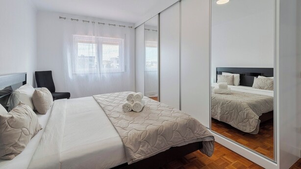Un dormitorio con cama, una silla, un espejo grande y una ventana con cortinas translúcidas.
