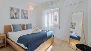 Une chambre à coucher moderne avec un lit en bois, une literie blanche et des touches de couleur bleue. On y trouve une fenêtre avec des rideaux, un climatiseur fixé au mur et des œuvres d’art au-dessus du lit.