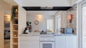 Une cuisine moderne avec des meubles blancs, un plan de travail en marbre et un four encastré. On y trouve un petit coin repas avec des chaises et une table, ainsi qu’une horloge au mur.