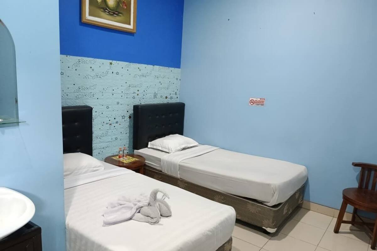 A hotel room with two single beds, a small table with a bottle, a chair, and a sink.