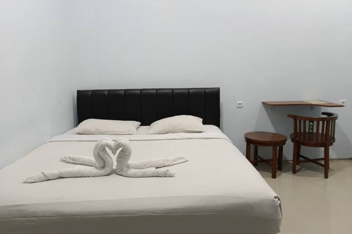 A single bed with a black headboard, two pillows, and a heart-shaped towel arrangement. There is a small wooden table and a chair to the right.