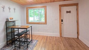 A room with a wooden ceiling, a window with a view of greenery, a door with a handle, and a table with chairs. - James Street Home Downtown Ludington (Ludington)