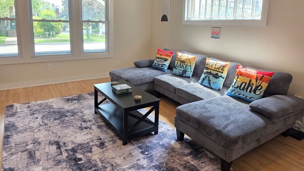 A living room with a sectional sofa, a coffee table, and a rug. There are large windows with a view of the outside. - James Street Home Downtown Ludington (Ludington)