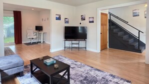 A modern living room with a flat-screen TV, a coffee table, and a sofa. There is a staircase leading to another level. - James Street Home Downtown Ludington (Ludington)