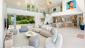 A spacious living area with a large sofa, a coffee table, and a dining area. There is a staircase leading to an upper level, and a kitchen area is visible in the background.