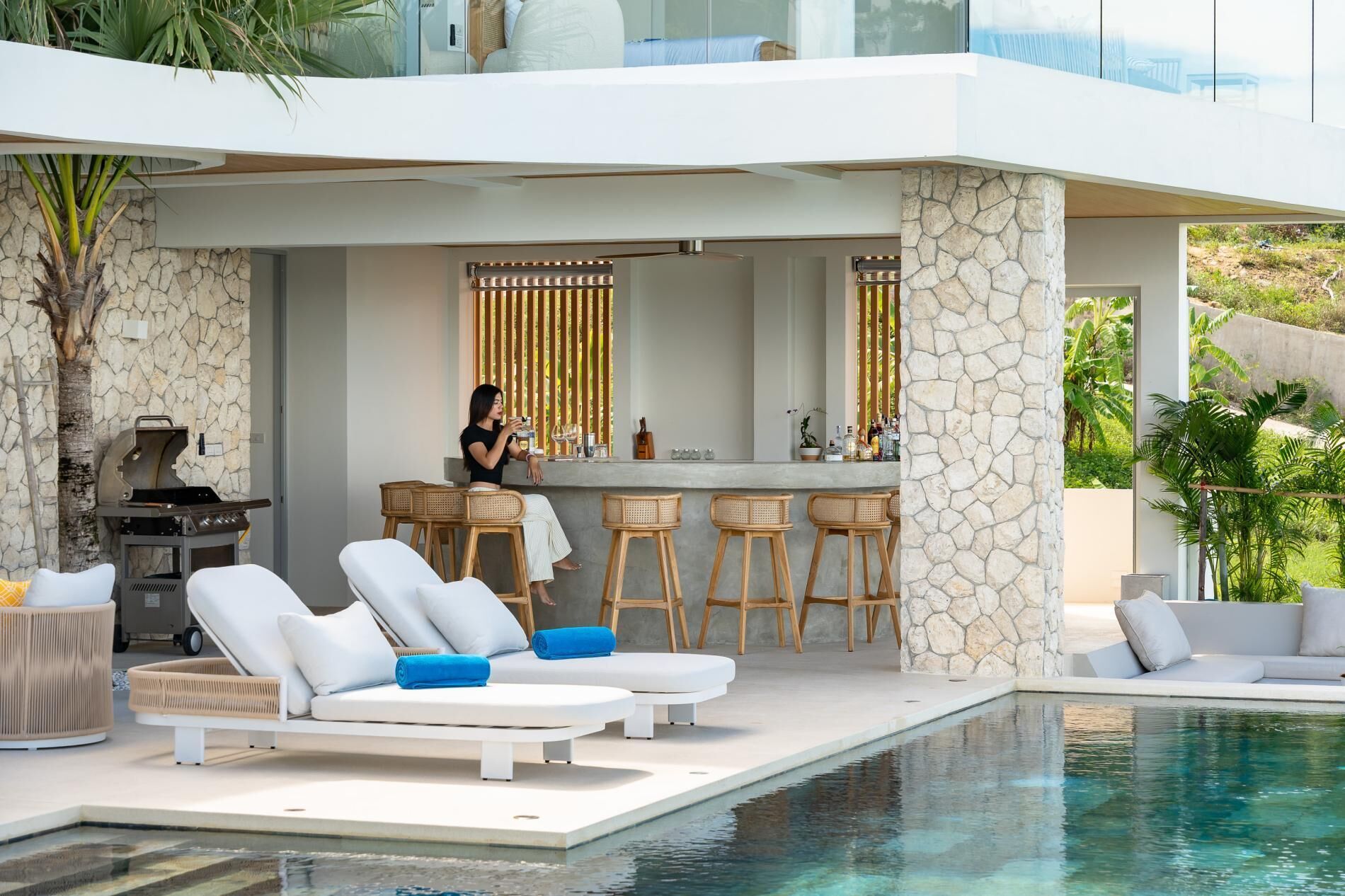 Un espace piscine extérieur moderne, doté de chaises longues, d’un bar et d’un barbecue.