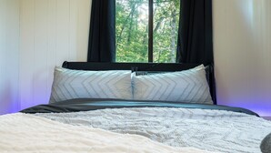 Una cama con colcha blanca, dos almohadas estampadas con un diseño de espiga, y vistas a árboles a través de una ventana con cortinas negras.