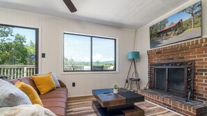 Una sala de estar con un sofá de cuero marrón, una mesa de centro de madera, una chimenea y grandes ventanas que ofrecen vistas a árboles y colinas.