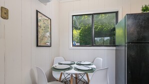 Comedor con una mesa preparada para cuatro, sillas blancas, un refrigerador negro y una ventana con vista a árboles.