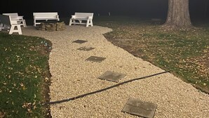 Un sendero de grava que conduce a bancos blancos bajo un árbol, con luces colgantes arriba.