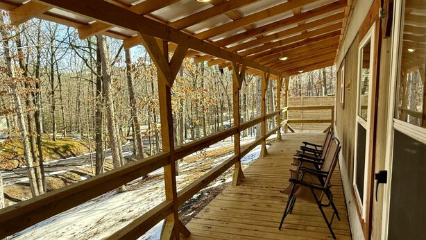 Une terrasse en bois couverte, dotée de bancs, qui offre une vue sur une zone boisée parsemée de plaques de neige.