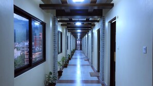 A corridor with a tiled floor, potted plants, and a view of buildings through large windows. - Hotel Queens Park (Pokhara)